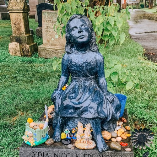 A blue statue of a girl sitting on a grave, surrounded by colorful toys, rocks, and a small blue container.