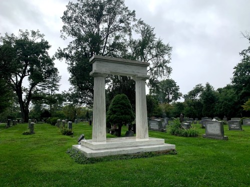 A stone monument with columns stands in a grassy cemetery surrounded by gravestones and trees under a cloudy sky.
