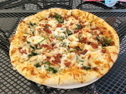 A freshly baked pizza topped with chicken, spinach, and bacon on a golden crust, served on a black mesh table.