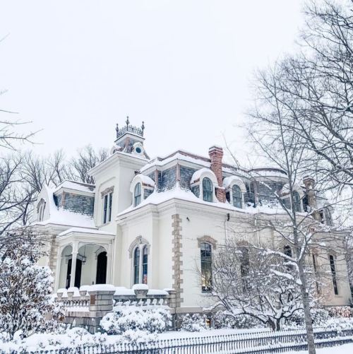 A grand, snow-covered mansion with intricate architecture and a turret, surrounded by bare trees in winter.
