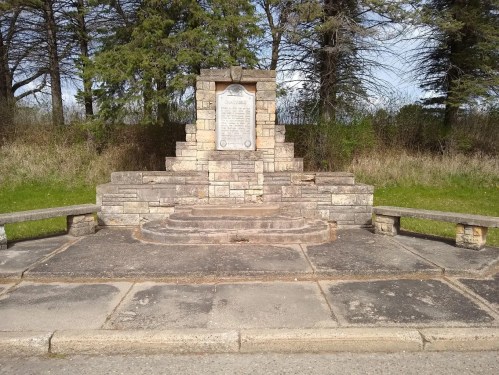 A stone monument with a plaque, surrounded by grass and trees, featuring steps and benches on either side.
