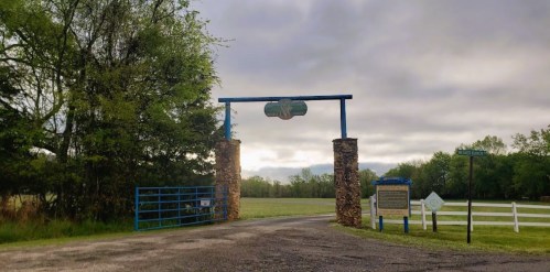 A stone entrance gate with blue accents leads to a grassy field under a cloudy sky. Signage is visible nearby.