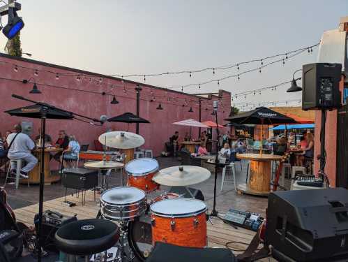 A lively outdoor venue with a stage set for a band, featuring tables, umbrellas, and people enjoying the atmosphere.