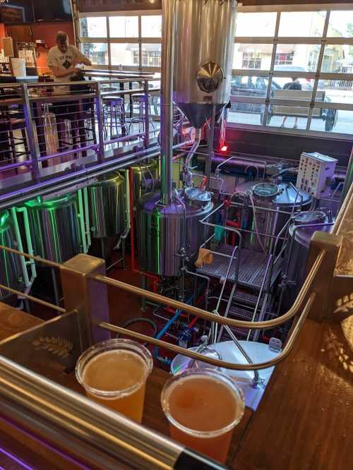 A brewery interior with brewing equipment and two cups of beer on a wooden ledge. A person is visible above.