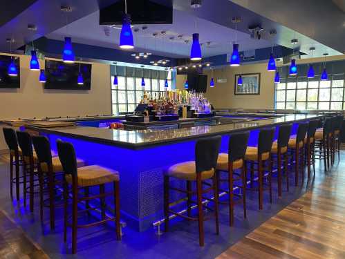 A modern bar with blue lighting, high stools, and a well-stocked counter, featuring multiple TVs and a spacious layout.