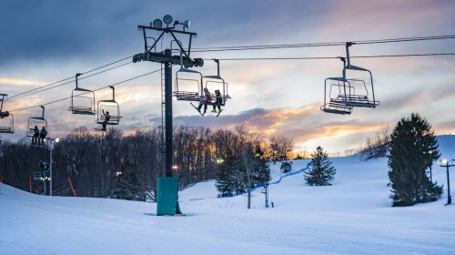Ski lift chairs against a sunset sky, with snow-covered slopes and trees in the background.