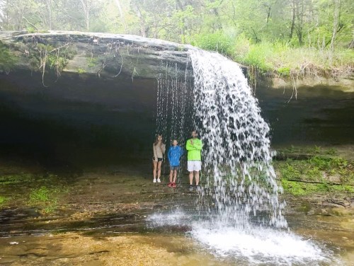 Three people stand under a waterfall, surrounded by lush greenery and rocky terrain. Water cascades down behind them.