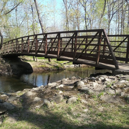 A wooden bridge arches over a calm stream, surrounded by trees and rocky banks in a sunny park setting.