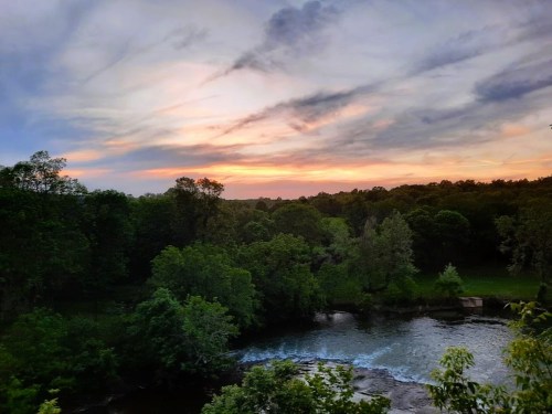 A serene sunset over a river, surrounded by lush green trees and a colorful sky.