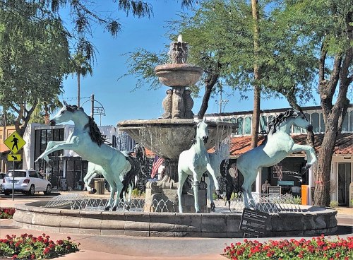 A fountain featuring three rearing horses, surrounded by greenery and flowers in a sunny plaza.