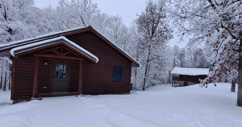 A cozy wooden cabin surrounded by snow-covered trees and a second building in a winter landscape.