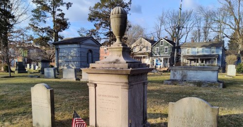 A cemetery scene featuring a prominent monument with an urn, surrounded by gravestones and trees in the background.