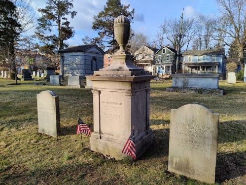 A historic grave marker with an urn, surrounded by smaller headstones and American flags in a cemetery.