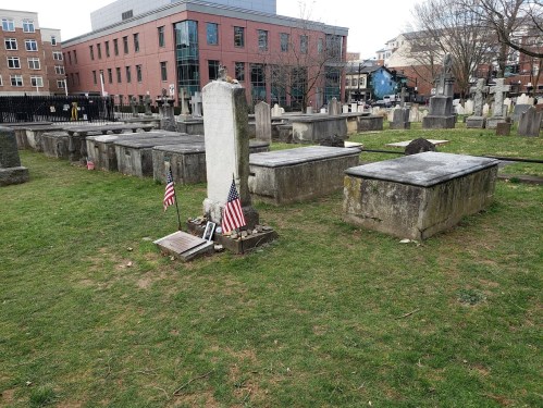 Graveyard with tombstones and small American flags, set against a backdrop of modern buildings and bare trees.
