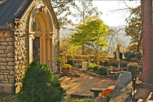 A stone building with an arched entrance surrounded by colorful flowers and trees in a serene garden setting.