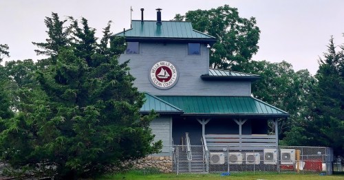 A gray building with a green roof and a circular sign, surrounded by trees and air conditioning units.
