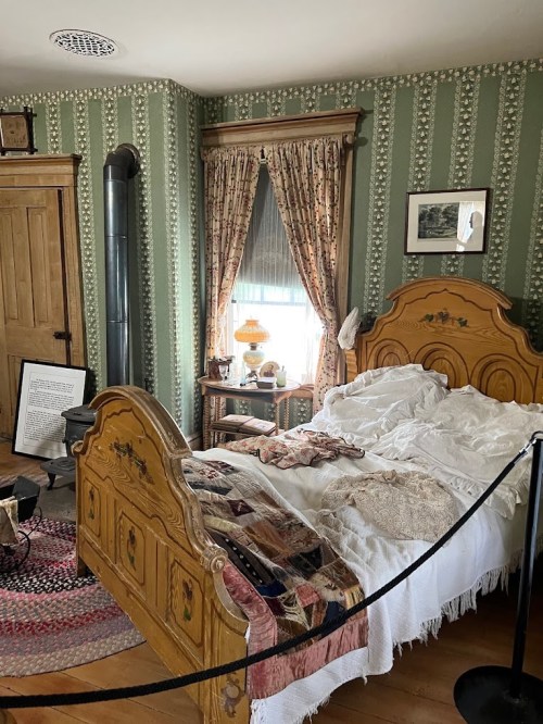 Cozy bedroom with a wooden bed, patterned wallpaper, and vintage decor, featuring a quilt and a window with curtains.