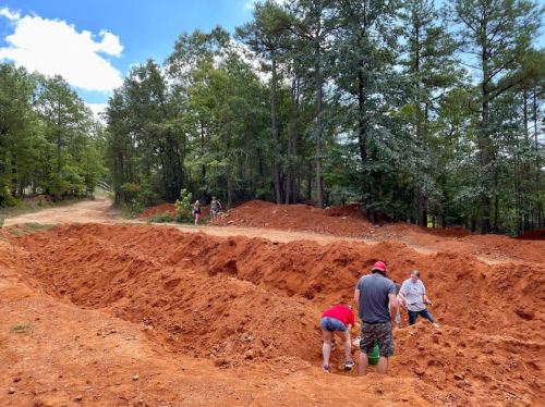 Workers digging in a large, red dirt trench surrounded by trees and a clear blue sky.