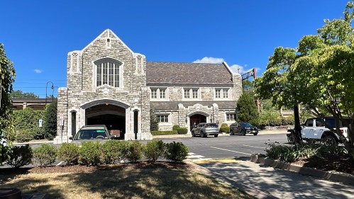 A stone building with a peaked roof, surrounded by trees and parked cars, under a clear blue sky.