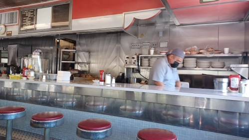 A diner interior with a counter, red stools, and a masked cook preparing food in a stainless steel kitchen.