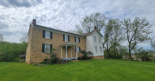 A two-story house with a stone exterior and white siding, surrounded by green grass and trees under a cloudy sky.