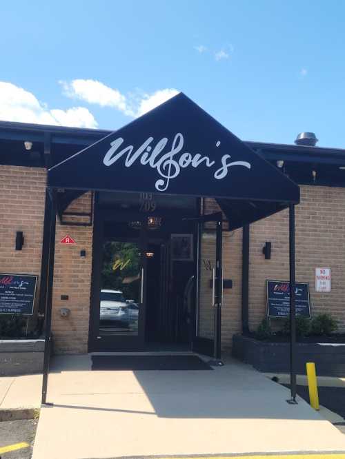 Entrance of Wilbon's restaurant with a black awning and musical note logo, surrounded by blue sky and greenery.