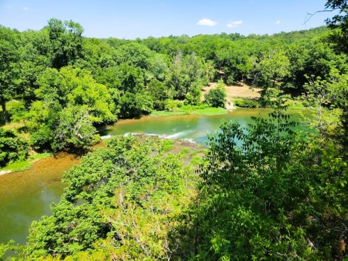 A serene river flows through lush green trees under a clear blue sky, with gentle rapids visible in the water.