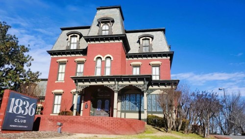Historic red brick mansion with a gray roof, featuring ornate details and a sign for the 1836 Club in front.