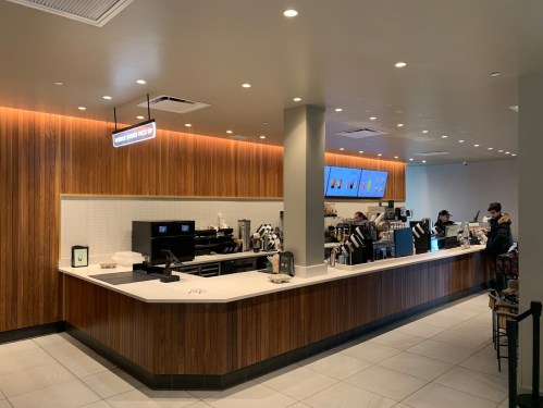 Modern coffee shop interior with a wooden counter, menu screens, and customers ordering at the counter.