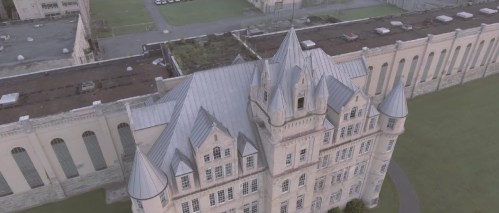 Aerial view of a large, historic building with a pointed roof and surrounding green space.