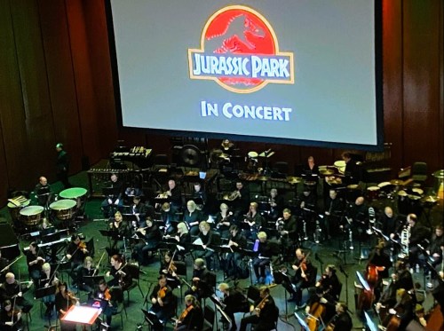 An orchestra performs with a large screen displaying "Jurassic Park In Concert" in the background.