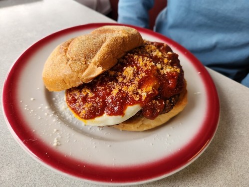 A sandwich on a plate with a soft bun, topped with a fried egg, chili, and sprinkled with cheese.