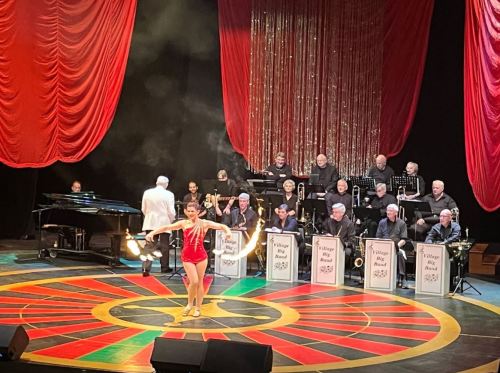 A performer in a red outfit juggles fire on stage, with a live band in the background and red curtains draping the scene.