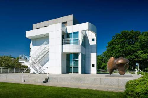 Modern white building with geometric design, featuring a staircase and a bronze sculpture in front, set against a clear blue sky.
