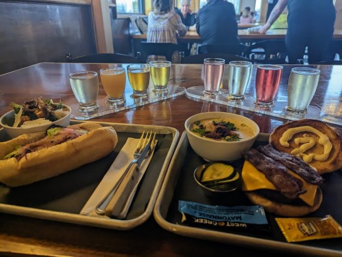 A table with a hot dog, sandwich, sides, and a flight of colorful drinks in small glasses.