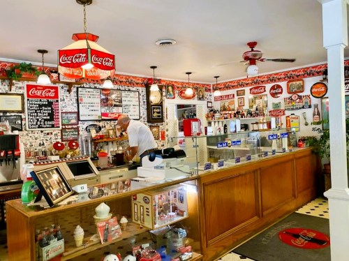 A vintage ice cream shop with a wooden counter, Coca-Cola decor, and a staff member serving customers.