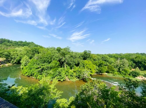 A serene river winding through lush green trees under a bright blue sky with wispy clouds.