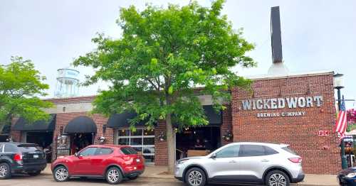 Exterior of Wicked Wort Brewing Company, featuring a brick facade, large windows, and parked cars under a green tree.