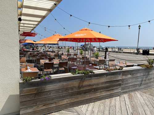 Outdoor dining area with wooden tables and orange umbrellas, overlooking a beach on a sunny day.