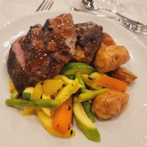 A plate of sliced beef with gravy, served alongside roasted potatoes and colorful sautéed vegetables.
