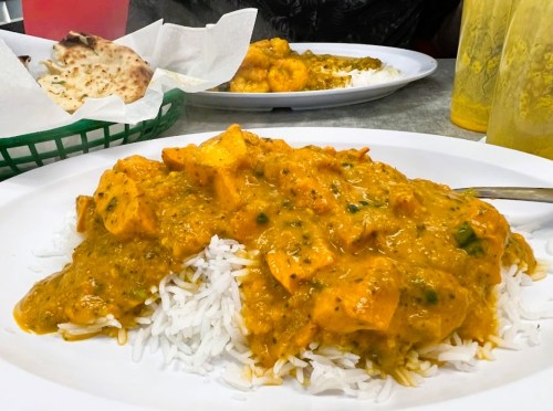 A plate of chicken curry served over white rice, with naan bread and drinks in the background.
