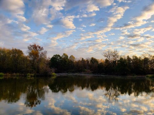 A serene lake reflects a colorful sky with clouds and trees, surrounded by autumn foliage.