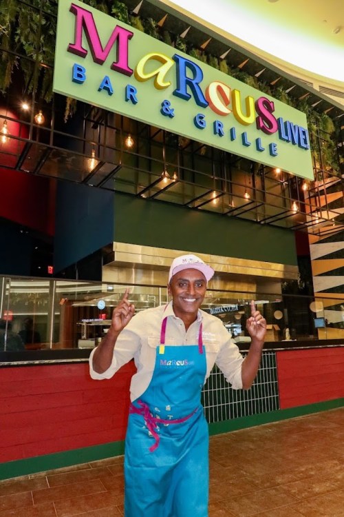 A smiling chef in an apron poses in front of the Marcus Live Bar & Grille sign, showcasing a vibrant restaurant interior.