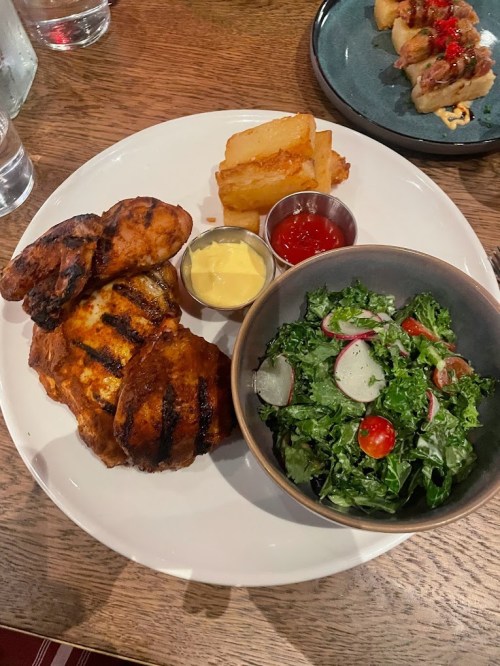 Grilled chicken, crispy potatoes, salad with radishes and tomatoes, served with dipping sauces on a white plate.