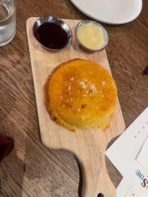 A round cornbread muffin on a wooden board, served with small bowls of jam and butter.