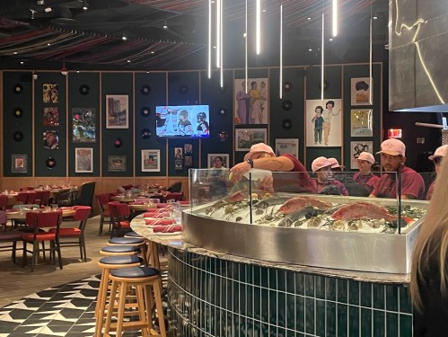 A vibrant restaurant interior with chefs preparing seafood at a display, colorful decor, and a TV in the background.