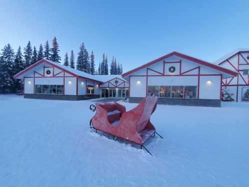 A red sleigh sits in front of a snowy building decorated for the holidays, surrounded by evergreen trees.