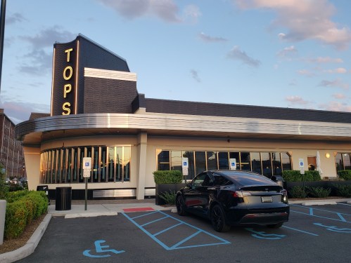 A black car parked in front of a retro diner named "Tops" with a modern design and large windows.