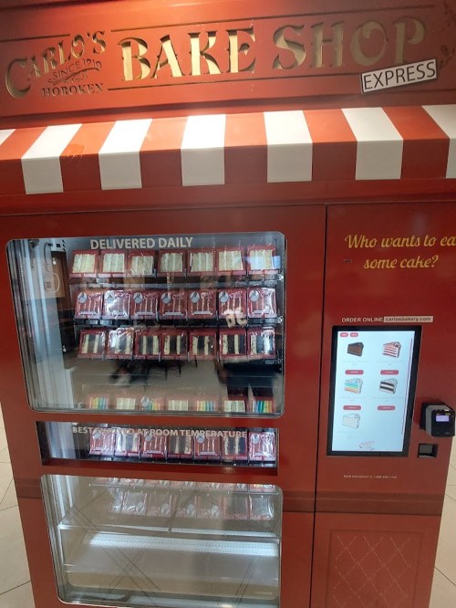 A red vending machine labeled "Carlo's Bake Shop" filled with cakes, featuring a touchscreen for ordering.