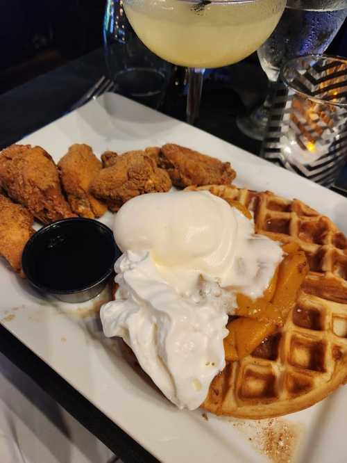 A plate featuring a waffle topped with ice cream and fruit, alongside fried chicken and a small cup of syrup.